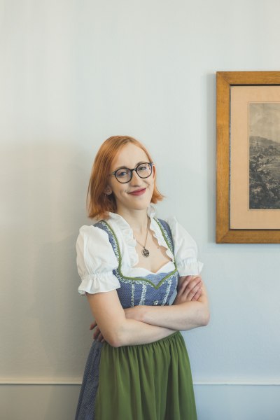 Im Service: Magdalena Bauer, © Niederösterreich Werbung/David Schreiber Eine Frau mit Brille und rotem Haar trägt ein traditionelles Dirndl und steht lächelnd vor einer weißen Wand neben einem gerahmten Bild.