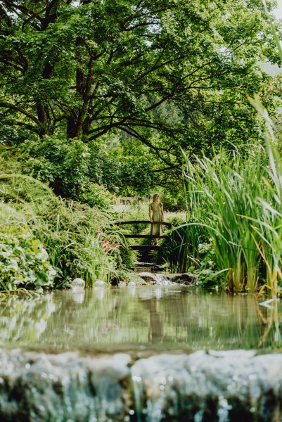 Die Deichgartenanlage ist von der Natur selbst geformt, © Niederösterreich Werbung/Rita Newman Eine Frau steht auf einer kleinen Brücke in einem üppigen, grünen Garten mit einem Teich im Vordergrund.