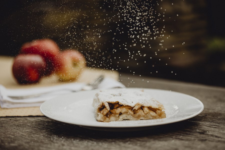 Hausgemachter Apfelstrudel, © Niederösterreich Werbung/Sophie Menegaldo Apfelstrudel mit Puderzucker auf Teller, Äpfel im Hintergrund.