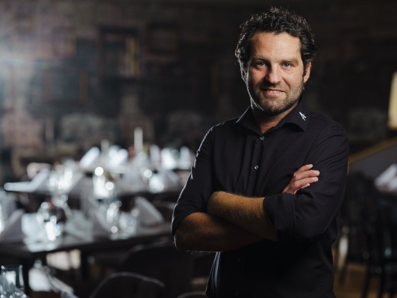 Showkochkurse mit Wirt Alexander Muhr, © Niederösterreich Werbung/Michael Reidinger Mann in schwarzem Hemd steht in elegantem Restaurant, Tische im Hintergrund.