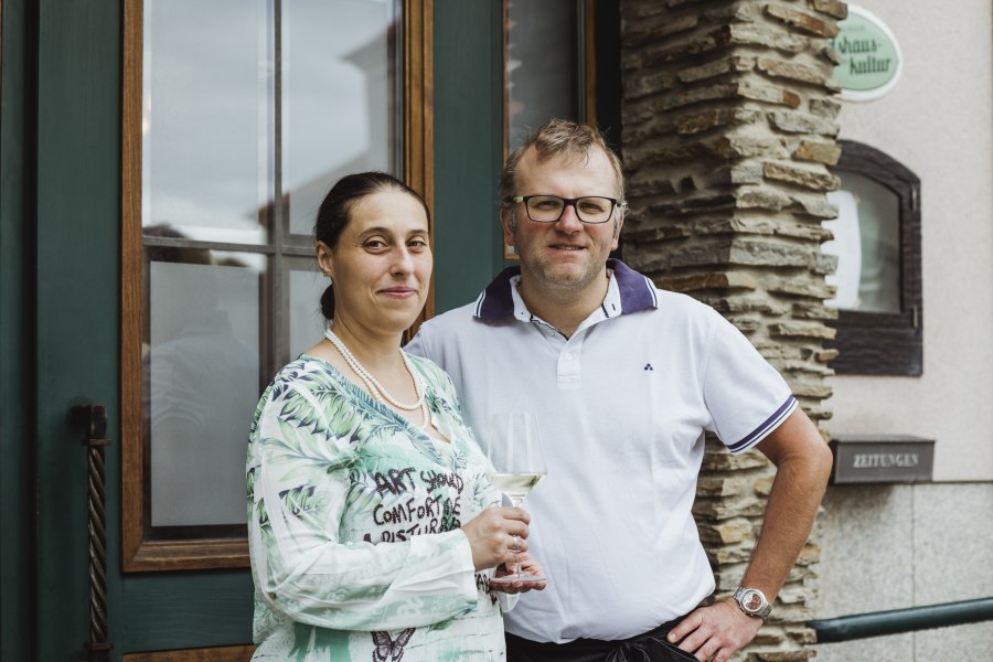 Wirtsleute Martina und Helmut Haas, © Niederösterreich Werbung/David Schreiber Ein Mann und eine Frau stehen lächelnd vor einem Gebäude. Die Frau hält ein Weinglas in der Hand.