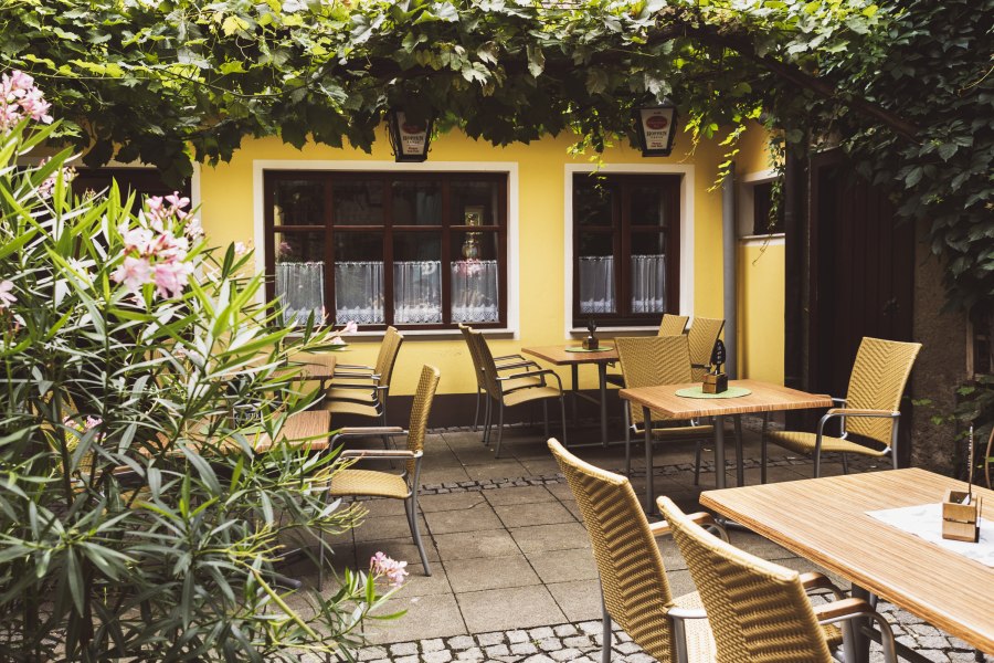 Ein typisch Weinviertler Wirtshaus eben, © Niederösterreich Werbung/David Schreiber Gemütlicher Gastgarten eines Wirtshauses mit gelber Fassade, Holztischen und Korbstühlen, umgeben von Pflanzen und Weinreben.
