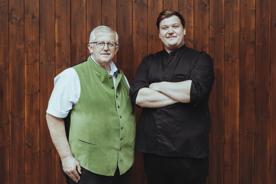 Reinhard Bürgmayr Junior und Senior, © Niederösterreich Werbung/David Schreiber Zwei Männer stehen vor einer Holzwand, einer trägt eine grüne Weste, der andere ein schwarzes Hemd.