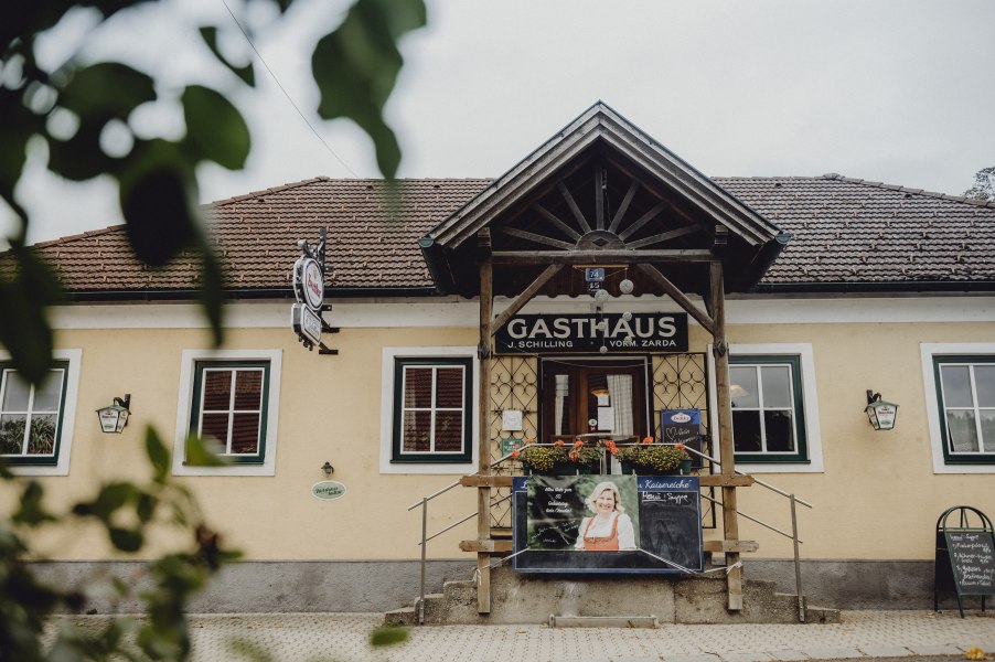 Wirtshaus mit Winter- und Gastgarten, © Niederösterreich Werbung/Rita Newman Eingang eines traditionellen Gasthauses mit Holzveranda und Werbetafel.