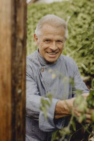 Gertrude Geidel, © Niederösterreich Werbung/Sophie Menegaldo Ein älterer Mann in einem blauen Hemd lächelt in die Kamera, umgeben von grünen Pflanzen.