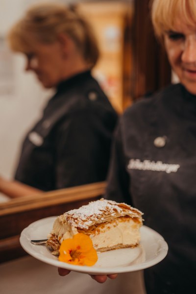 Selbstgemachte Mehlspeisen, © Niederösterreich Werbung/Daniela Führer Eine Person hält einen Teller mit einem Stück Torte, dekoriert mit einer Blume.