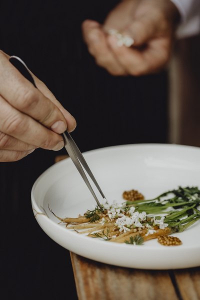 Neues bei Chef’s Table oder Triad privat, © Niederösterreich Werbung/Sophie Menegaldo Nahaufnahme eines Tellers mit kunstvoll angerichteten Gemüsen und Blüten, eine Hand arrangiert mit einer Pinzette.