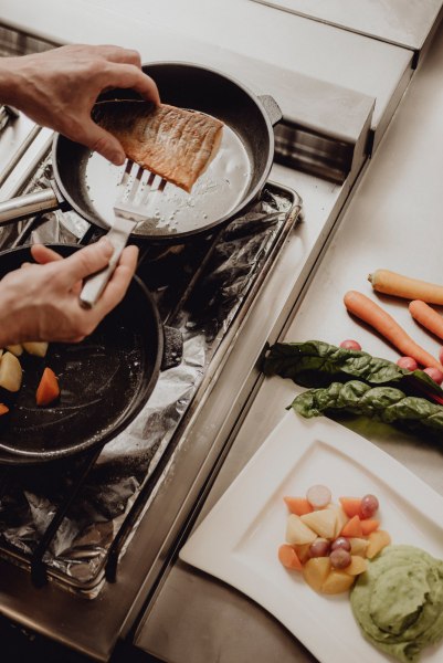 Die Küchenlinie: zuverlässige Bodenständigkeit mit frischen Ideen, © Niederösterreich Werbung/Rita Newman Eine Person brät ein Stück Fisch in einer Pfanne auf einem Herd. Daneben liegen Gemüse und ein Teller mit Beilagen.