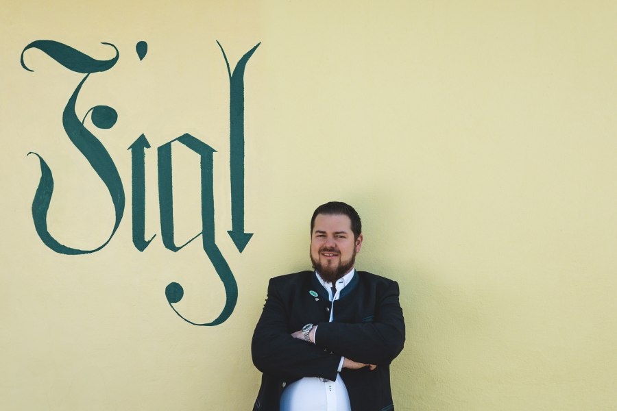 Matthias Strunz, der Wirt vom Figl, © Niederösterreich Werbung/David Schreiber Ein Mann in traditioneller Kleidung steht vor einer gelben Wand mit dem Schriftzug 'Figl'.