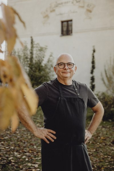 Wirt Manfred Tastl, © Niederösterreich Werbung/Sophie Menegaldo Mann mit Brille und Schürze steht lächelnd im herbstlichen Garten.