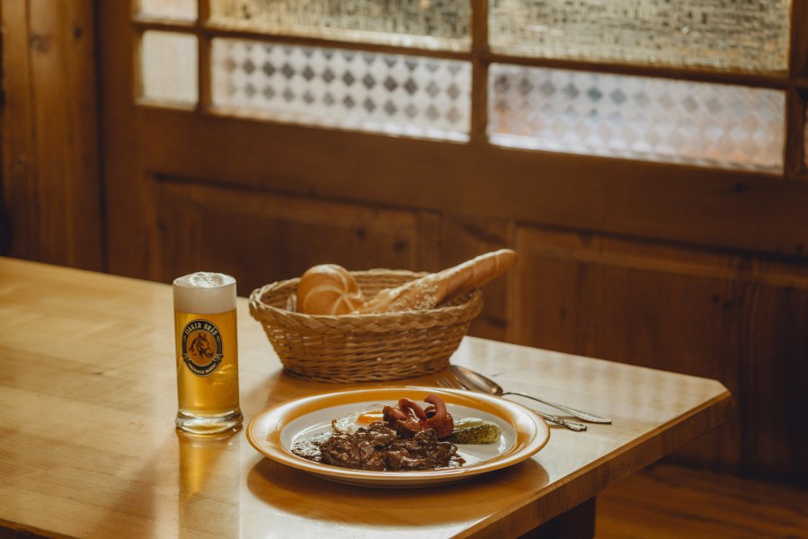 Zum Fiakergulasch passt das helle Fiakerbräu, © Niederösterreich Werbung/Rita Newman Ein Teller mit Fiakergulasch, Spiegelei und Gurke, daneben ein Glas Bier und ein Brotkorb auf einem Holztisch.