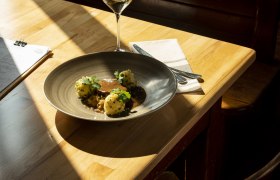Geschmorter Schweinebauch, Chilikraut, Erdäpfelknödel in knusperiger Schweineschwarte gewälzt, © Niederösterreich Werbung/Michael Reidinger Teller mit Fleisch, Knödeln und Soße, Weinglas auf Holztisch im Sonnenlicht.