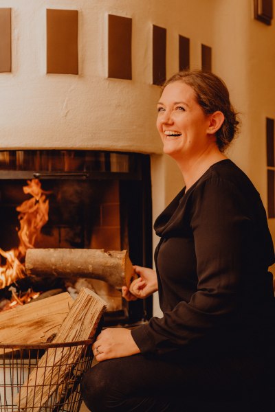 Wirtin Eva Maria Reisenbauer, © Niederösterreich Werbung/Klaus Engelmayer Frau lächelt vor einem Kamin mit brennendem Feuer.