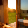 roon_blick-auf-terrasse, © Franz Atteneder Blick aus einem Zimmer auf eine Terrasse bei Sonnenuntergang, mit Wiese und Wald im Hintergrund.