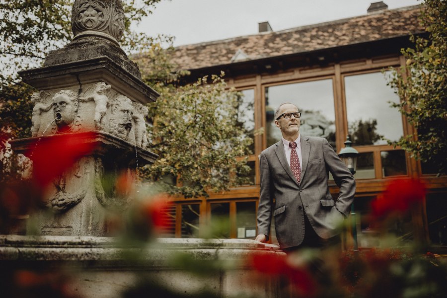 Andreas Horvath, © Niederösterreich Werbung/Sophie Menegaldo Ein Mann im Anzug steht neben einem steinernen Brunnen mit Wasserspeiern, umgeben von roten Blumen und einem Gebäude im Hintergrund.