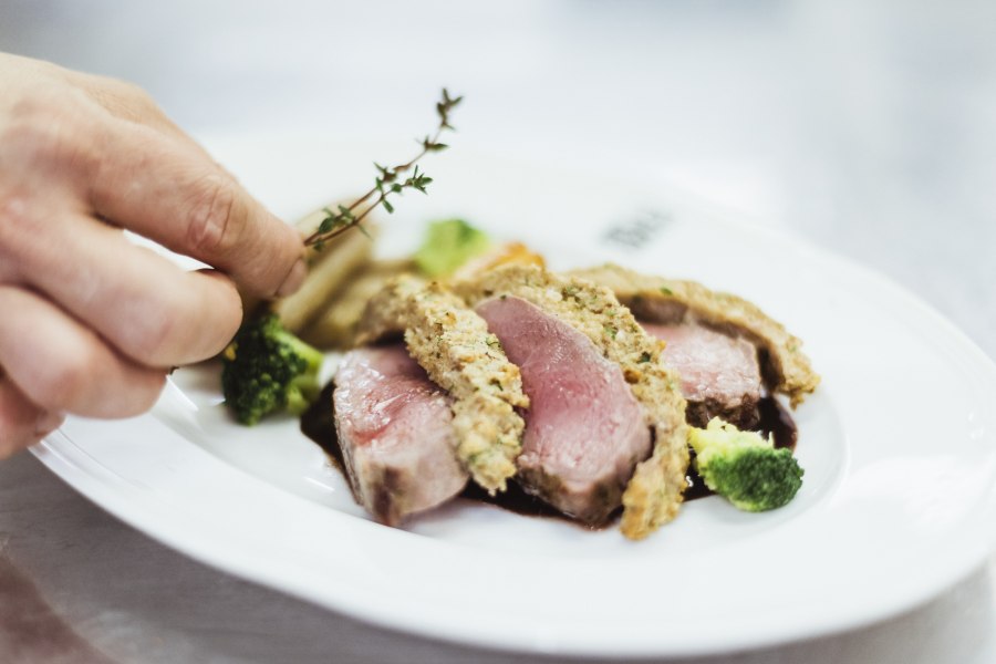 Rosa Hirschrücken mit Maronischupfnudeln, © Niederösterreich Werbung/David Schreiber Nahaufnahme eines rosa gebratenen Fleischgerichts mit Kräuterkruste, Brokkoli und einer Hand, die Thymian hinzufügt.