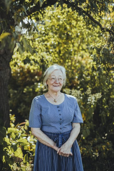 Wirtin Elisabeth Bauer, © Niederösterreich Werbung/David Schreiber Ältere Frau in blauem Kleid steht lächelnd im Garten.