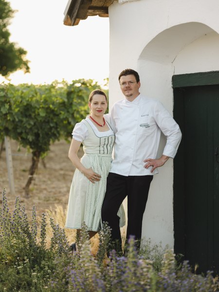 Wirtsleute Daniela und Harald Graf, © Niederösterreich Werbung/Michael Reidinger Ein Paar in traditioneller Kleidung steht vor einem weißen Gebäude neben einem Weinberg.