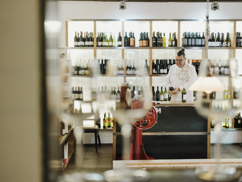 Mann in Vinothek mit Regalen voller Weinflaschen, unscharfe Gl&auml;ser im Vordergrund.