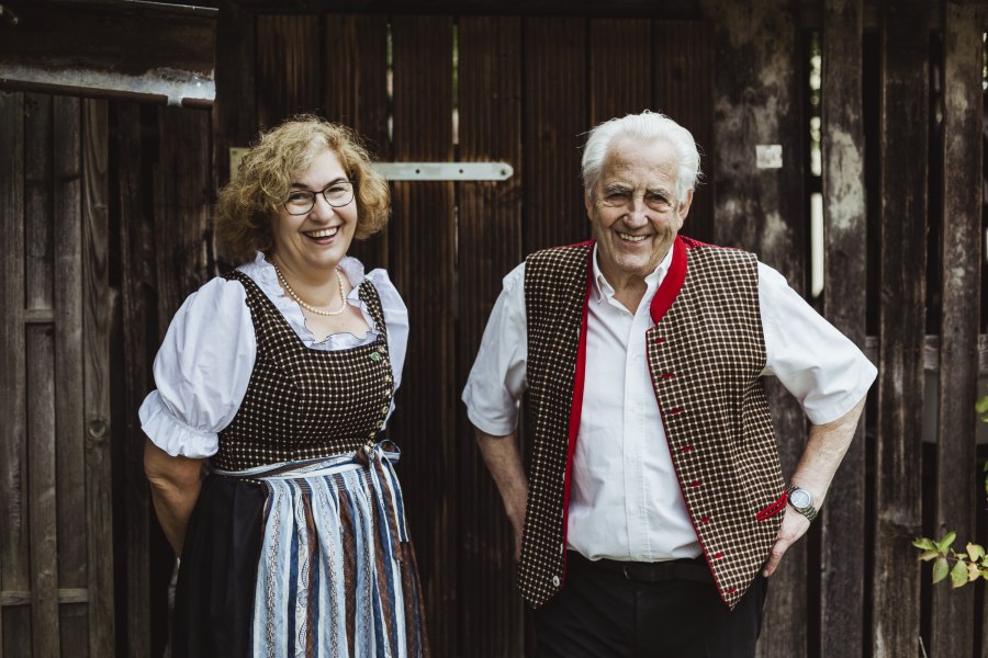 Zwei Personen in traditioneller Tracht vor einer Holzwand, l&auml;chelnd.