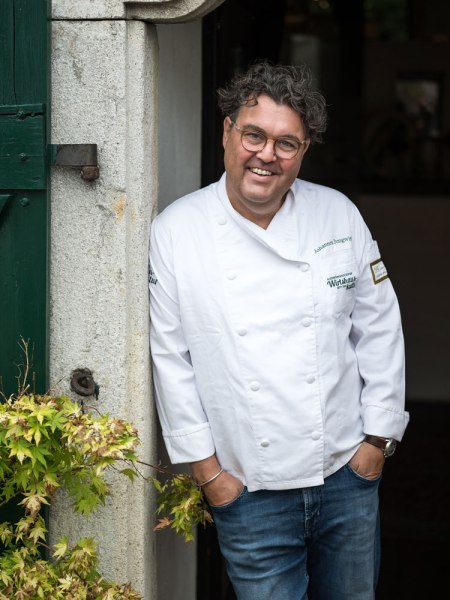 Mann in wei&szlig;er Kochjacke lehnt l&auml;chelnd an einer T&uuml;r.