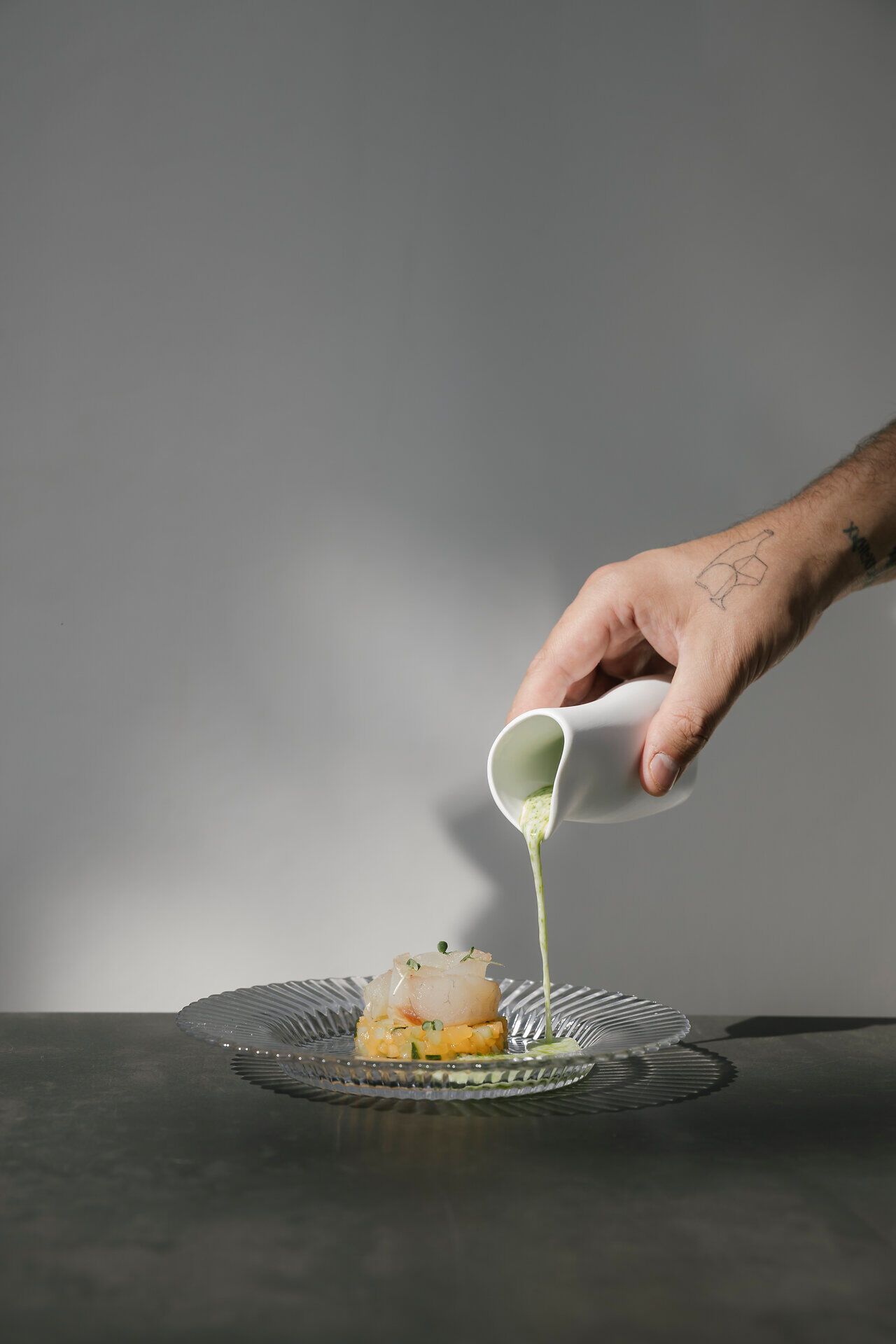 Eine Hand gießt eine grüne Sauce aus einem weißen Kännchen auf ein angerichtetes Gericht mit Fisch und klein geschnittenem Gemüse auf einem Glasteller.
