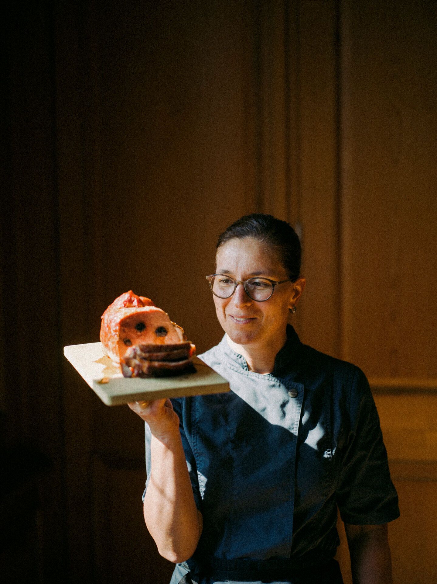 In einem warmen Lichtstrahl präsentiert eine Köchin stolz das köstliche Mostbratl, ein traditionelles Gericht der Region. Die Atmosphäre ist geprägt von herzlicher Gastfreundschaft und der Liebe zur regionalen Küche, die hier im Mostviertel zelebriert wird.