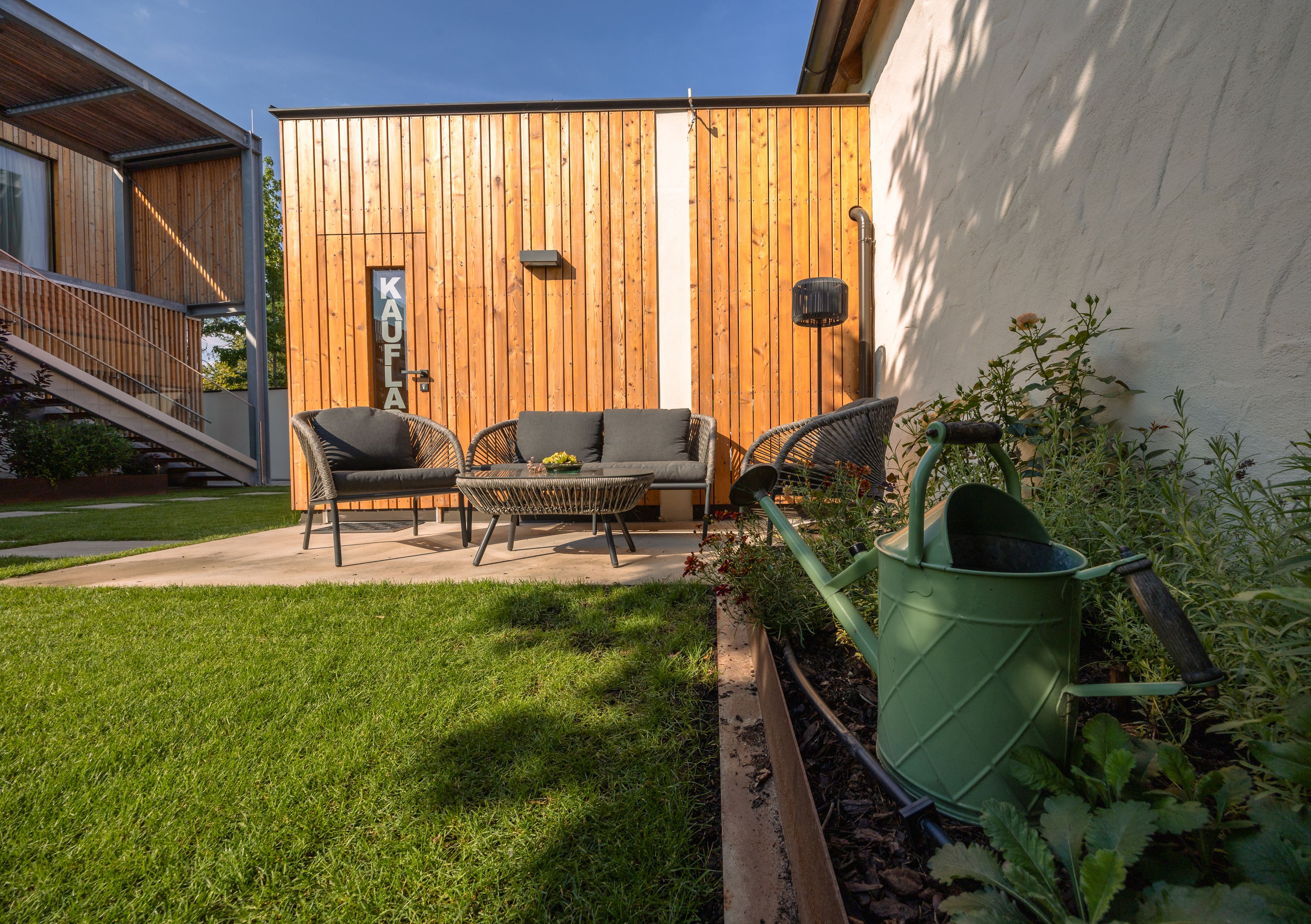 Garden area with seating furniture in front of a wooden house, watering can in the foreground.