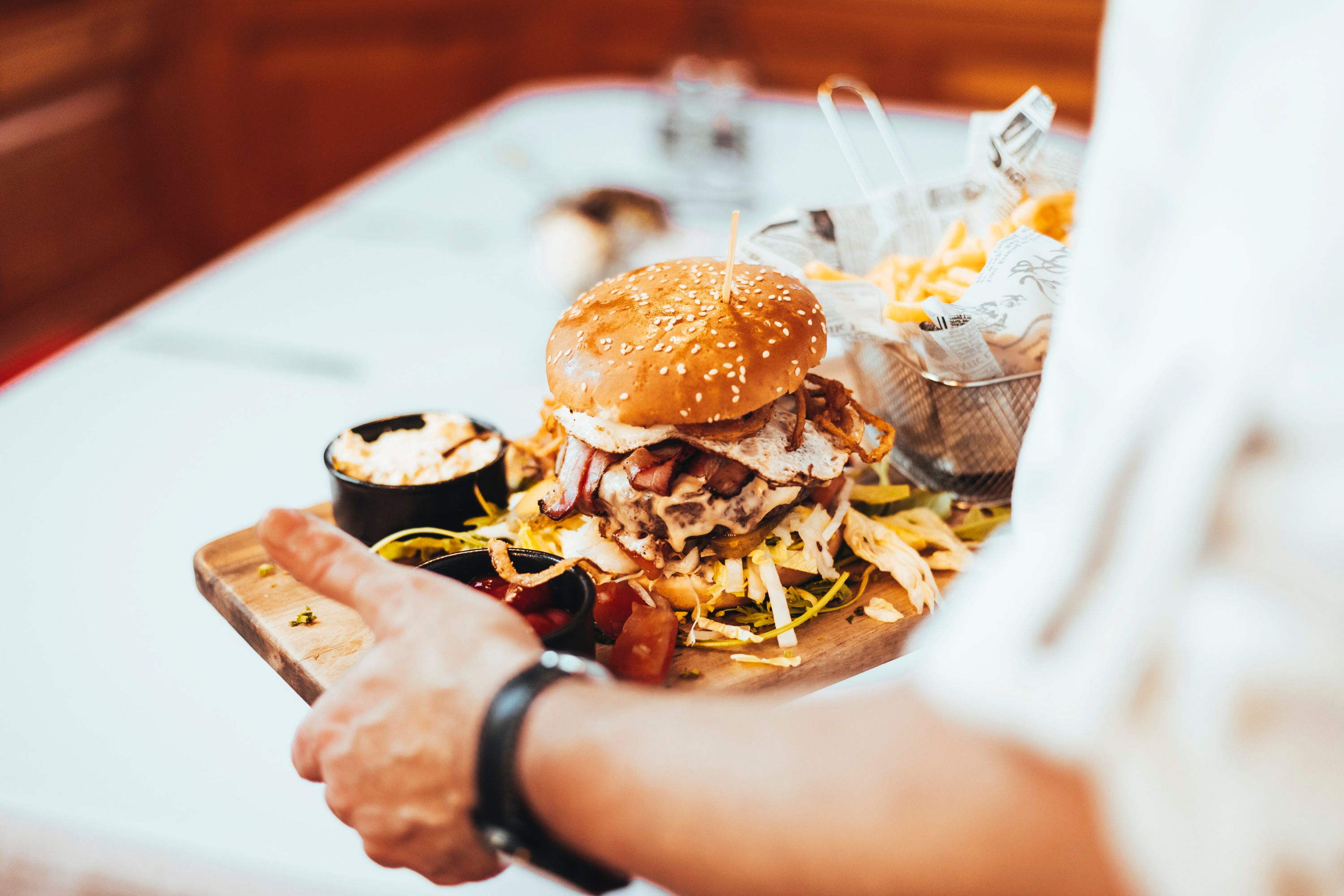 Ein Burger mit Sesambrötchen, Speck und Spiegelei auf einem Holzbrett, serviert mit Pommes und Dips.