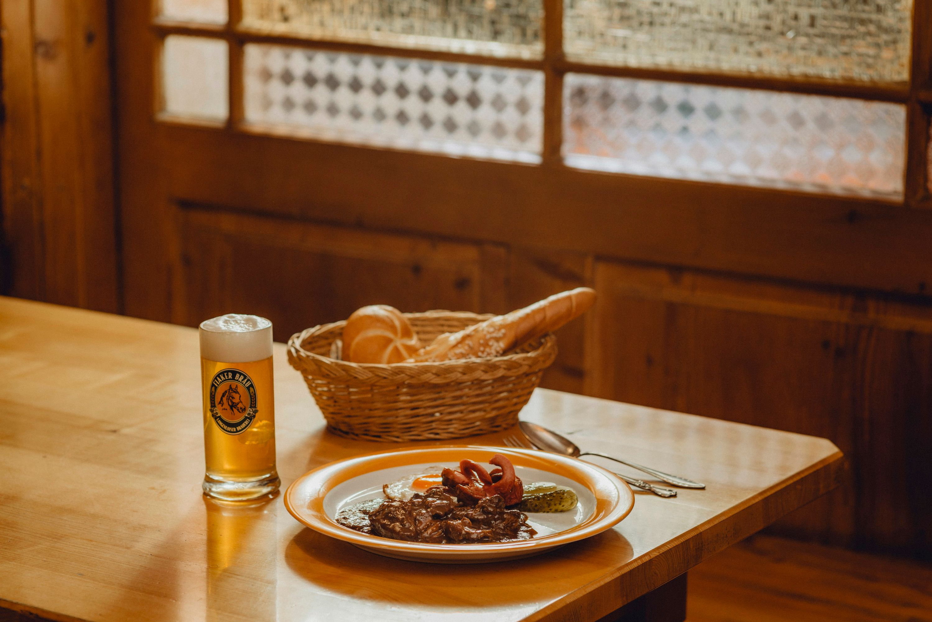 Ein Teller mit Fiakergulasch, Spiegelei und Gurke, daneben ein Glas Bier und ein Brotkorb auf einem Holztisch.