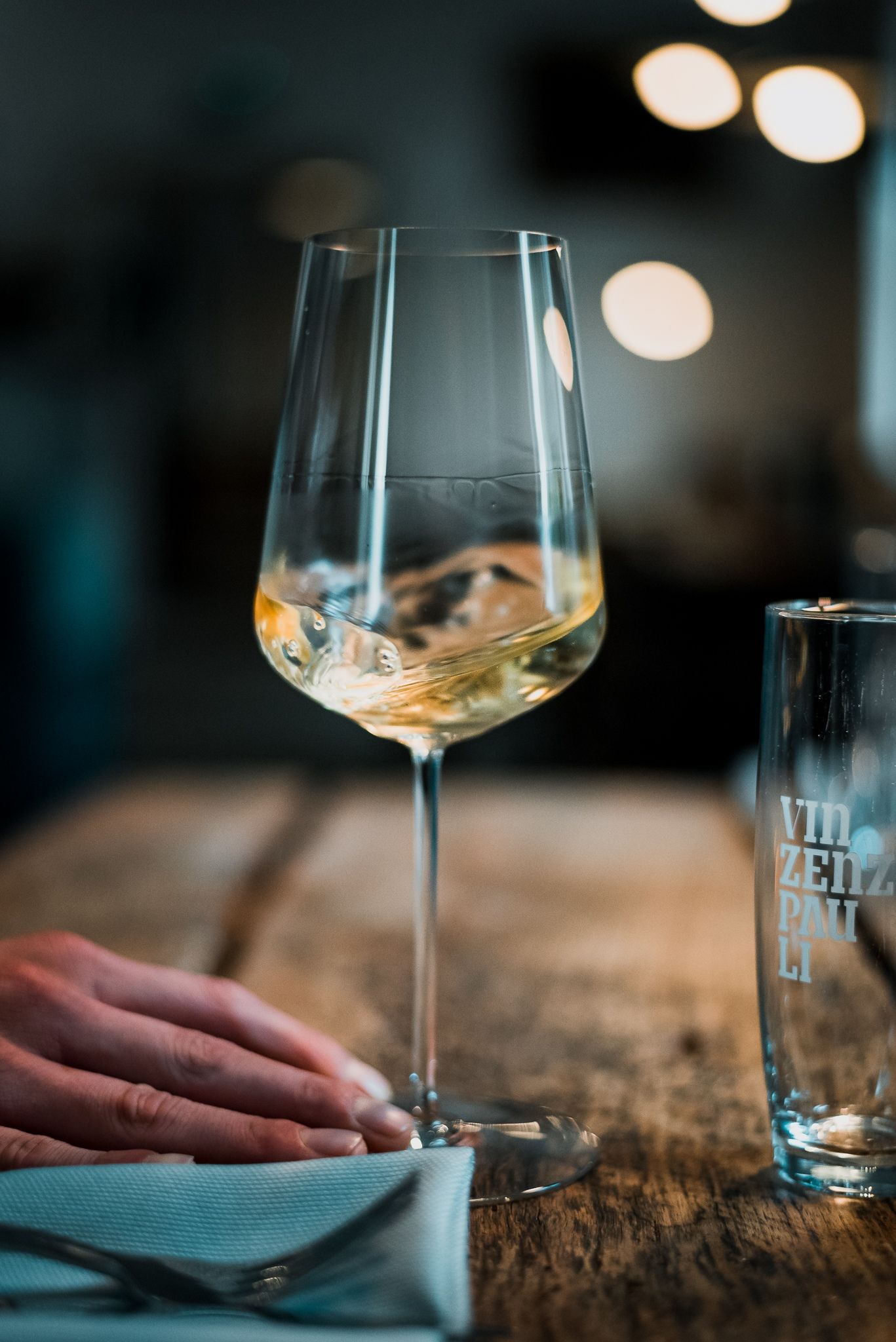 Ein Weinglas mit Weißwein auf einem Holztisch, daneben ein Glas mit der Aufschrift 'Vinzenz Pauli'.