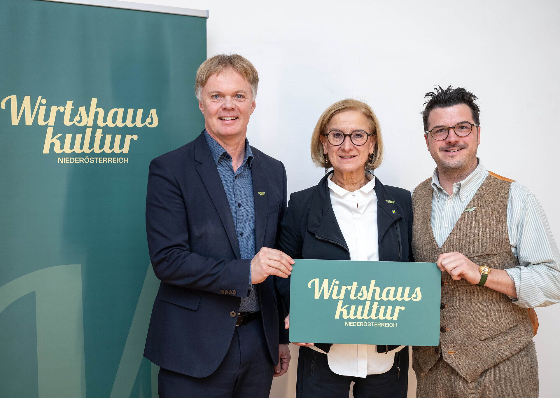 Drei Personen stehen vor einem Roll-up der Wirtshauskultur Niederösterreich und halten gemeinsam ein Schild mit dem Logo der Wirtshauskultur in die Kamera.