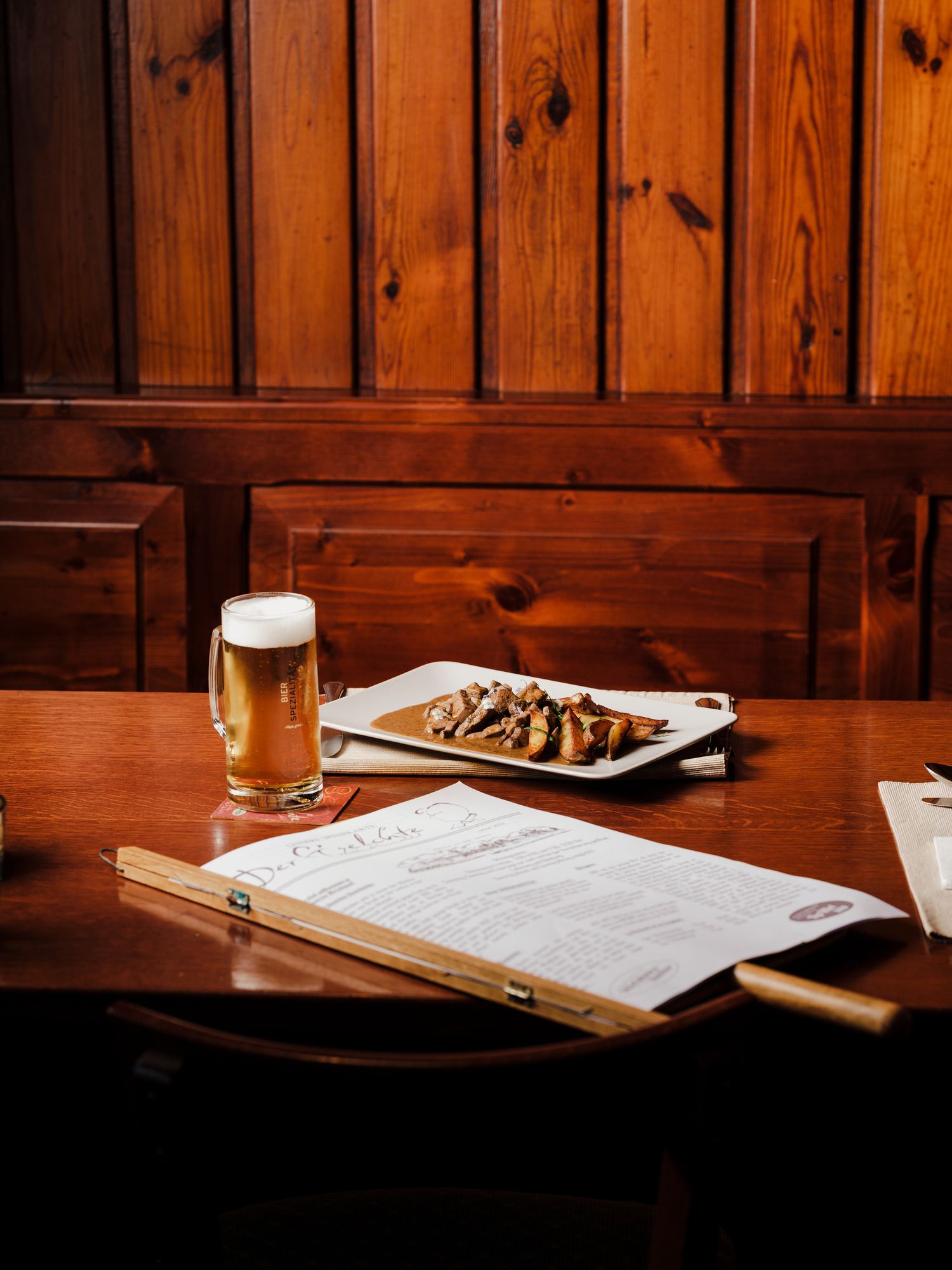 Holztisch mit Bier, Speisekarte und Teller mit Fleischgericht.