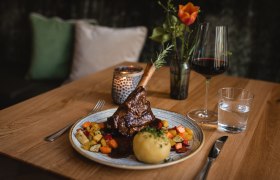 Ein Teller mit Lammstelze, Kn&ouml;del und Gem&uuml;se, daneben ein Glas Rotwein und Wasser auf einem Holztisch.