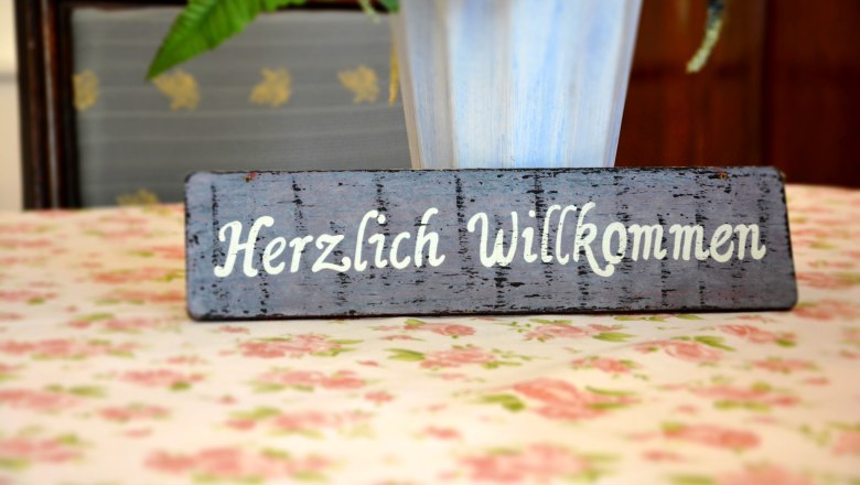 Ein Holzschild mit der Aufschrift 'Herzlich Willkommen' steht auf einem blumigen Tisch.