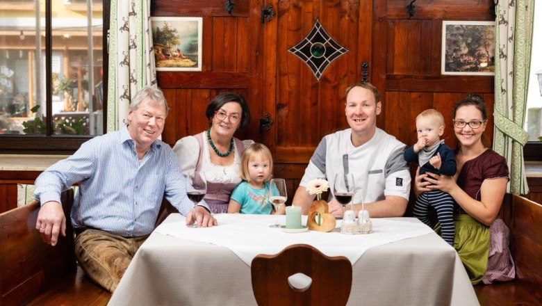 Familie sitzt an einem Tisch vor einer rustikalen Holzwand. Auf dem Tisch sind 3 Weingläser und Deko.