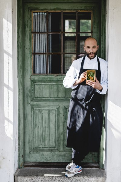 Ein Mann in Kochkleidung hält eine Schachtel mit Kräutern vor einer grünen Holztür.