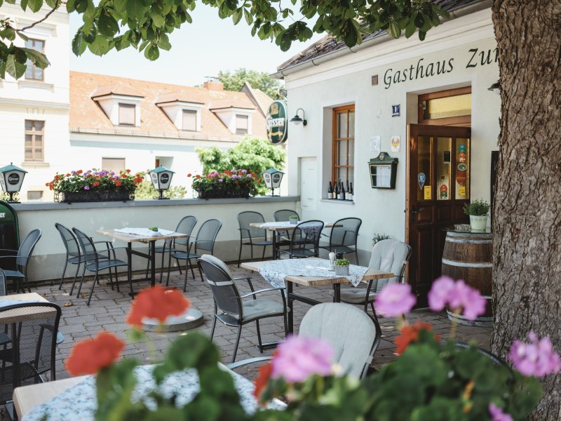 Schattiger Gastgarten mit Tischen und St&uuml;hlen vor einem Gasthaus.