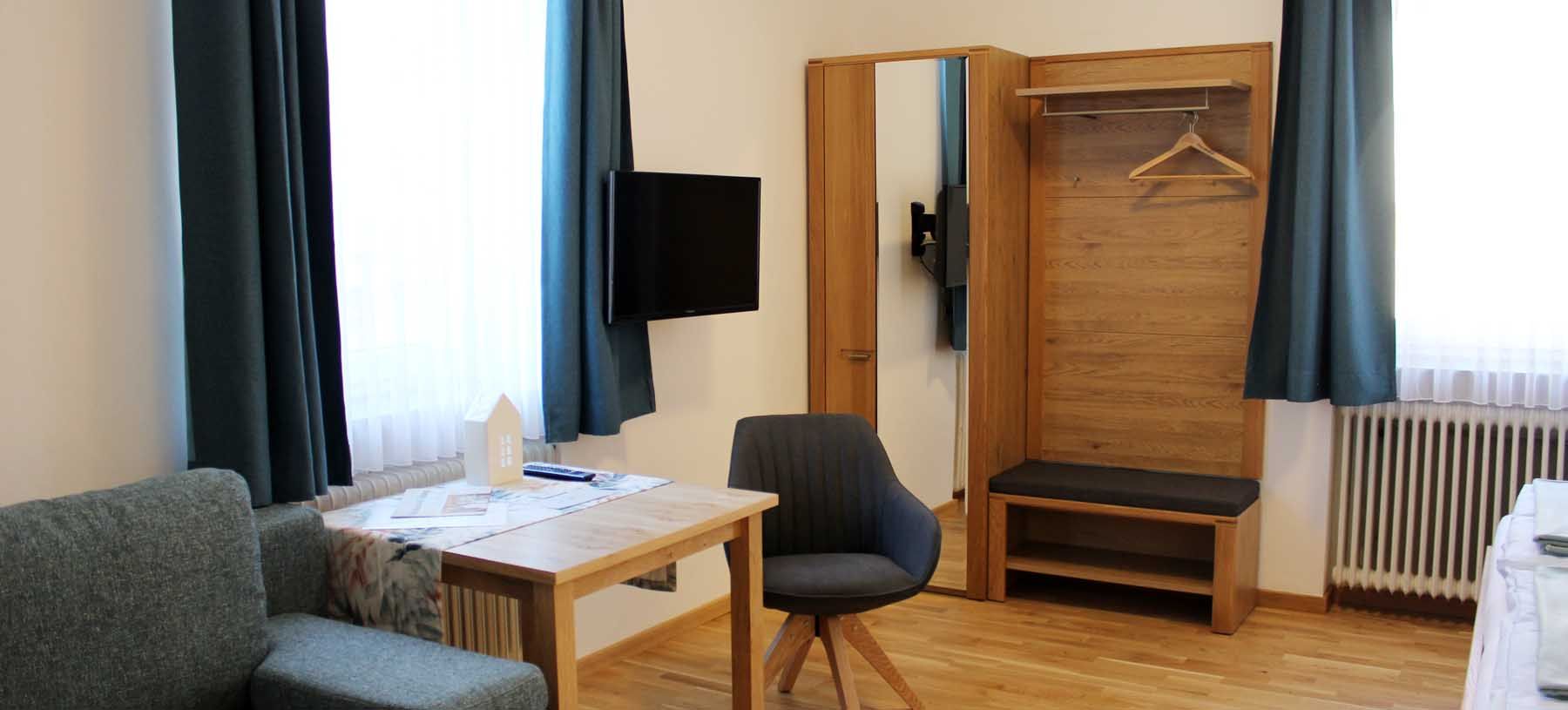 Modernes Gästezimmer mit Tisch, Stuhl, Kleiderschrank, Sofa, Fernseher und blauen Vorhängen.
