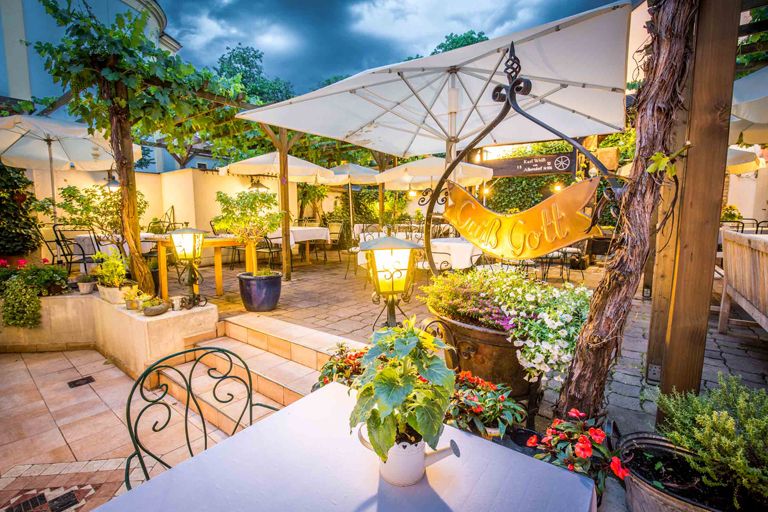 Gastgarten am Abend mit Beleuchtung und vielen Sitzmöglichkeiten, Pflanzen und Schirmen.