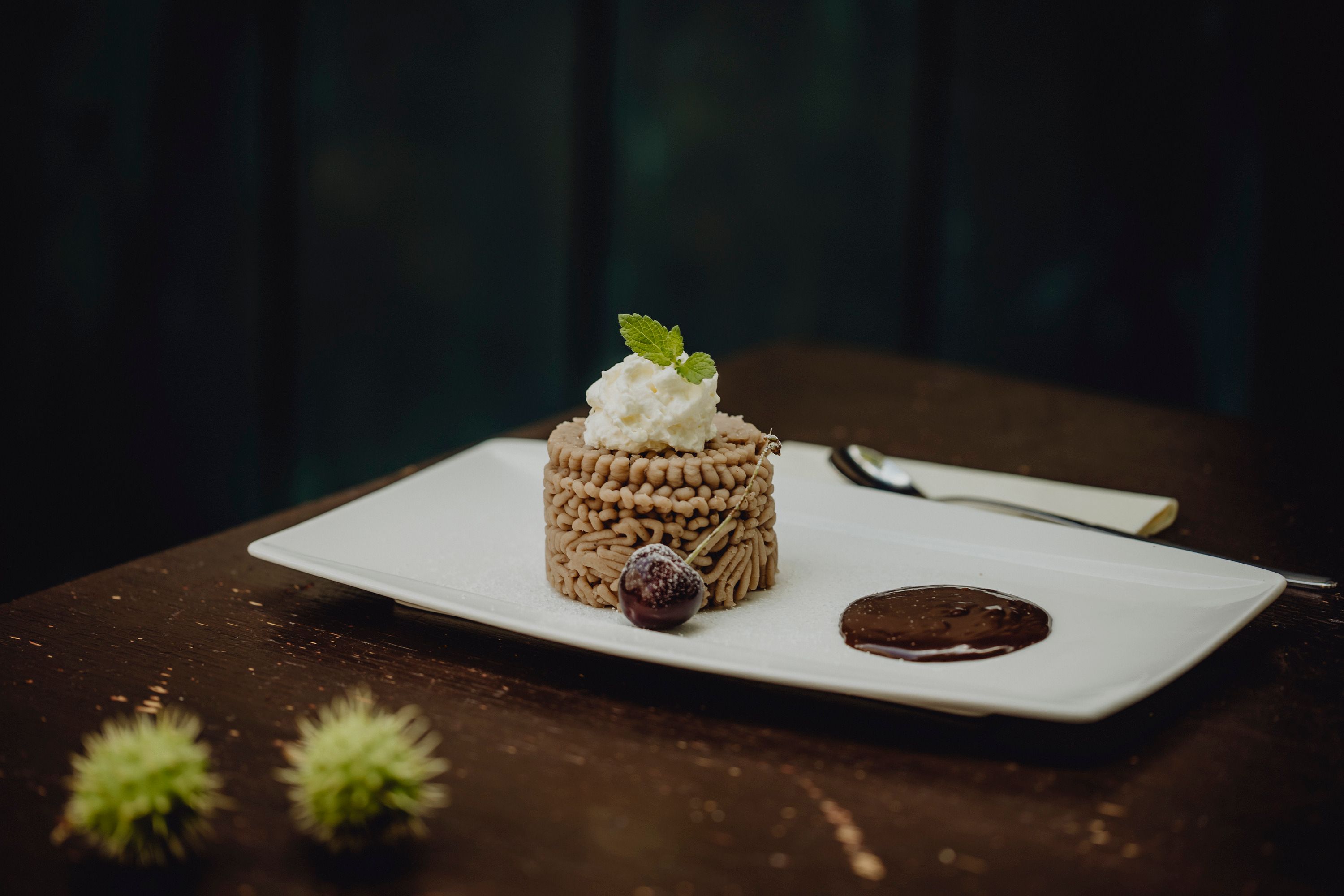 Ein Dessert aus Kastanienmousse auf einem weißen Teller, garniert mit Sahne und Minzblatt, daneben eine Schokoladensauce und eine Kirsche.