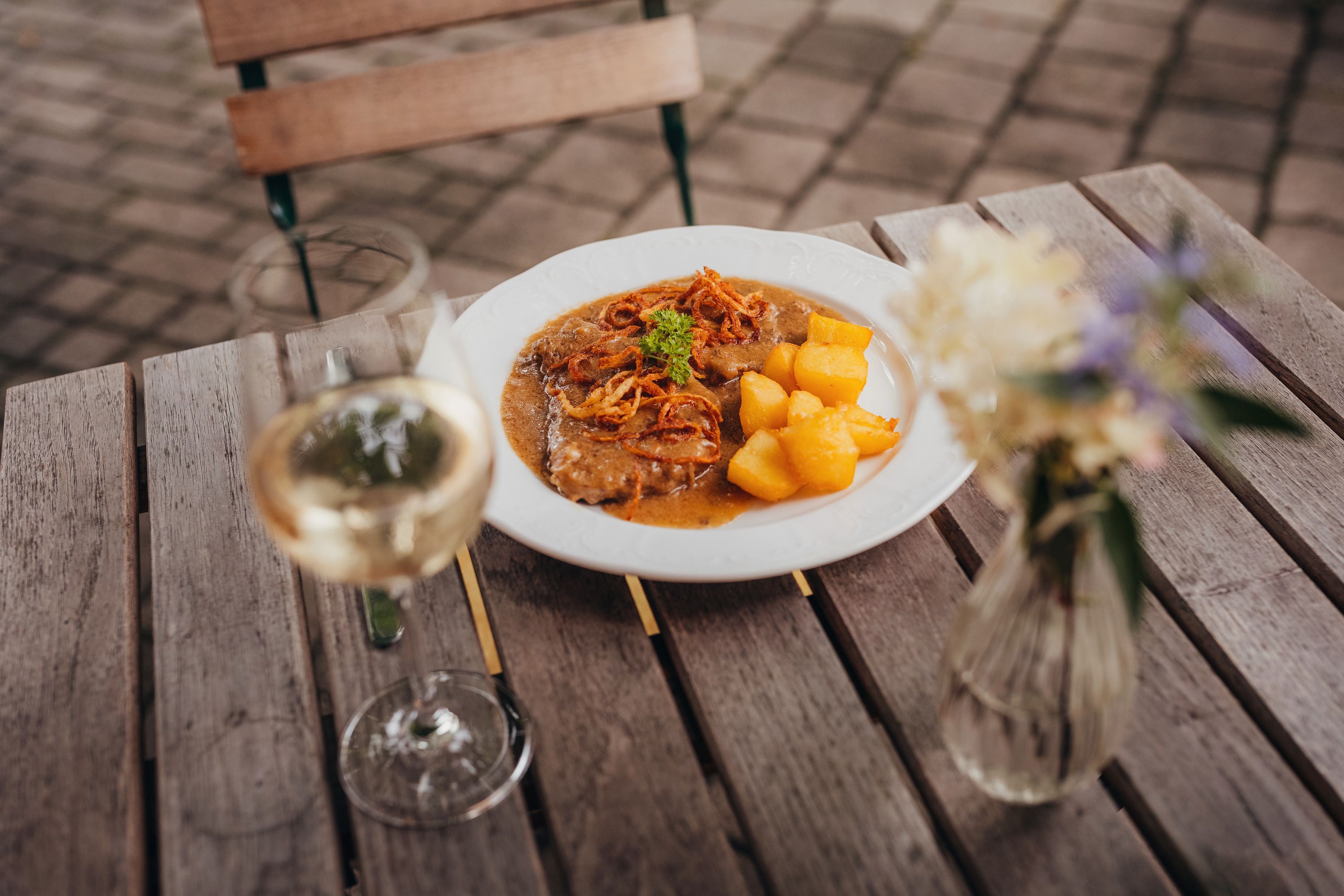 A plate of roast onions and roast potatoes on a wooden table, next to it a glass of white wine and a vase of flowers.