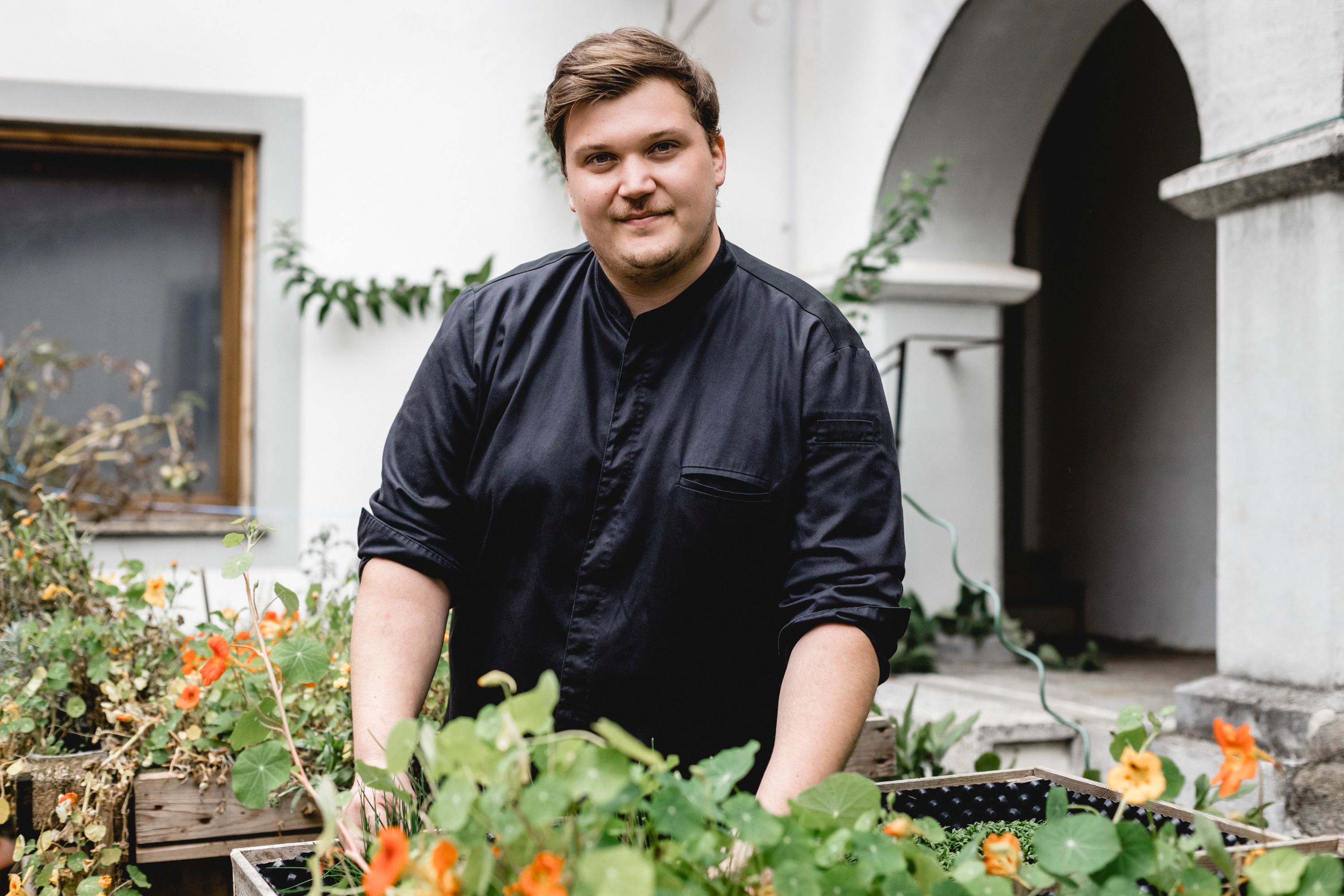 Ein Mann in schwarzer Kleidung steht in einem Garten mit Blumen und Pflanzen.