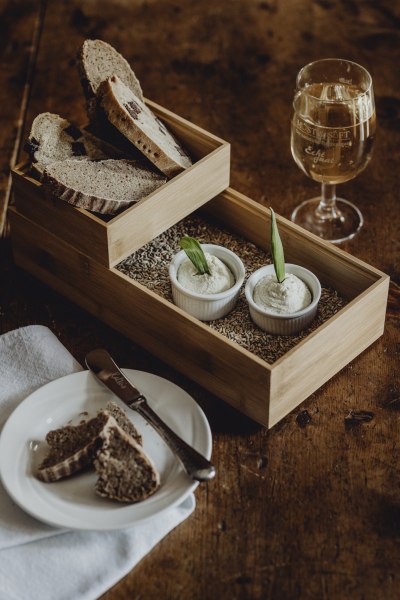 Ein Holztablett mit Brotscheiben, zwei Sch&auml;lchen mit Aufstrich und ein Glas Wei&szlig;wein auf einem Holztisch.