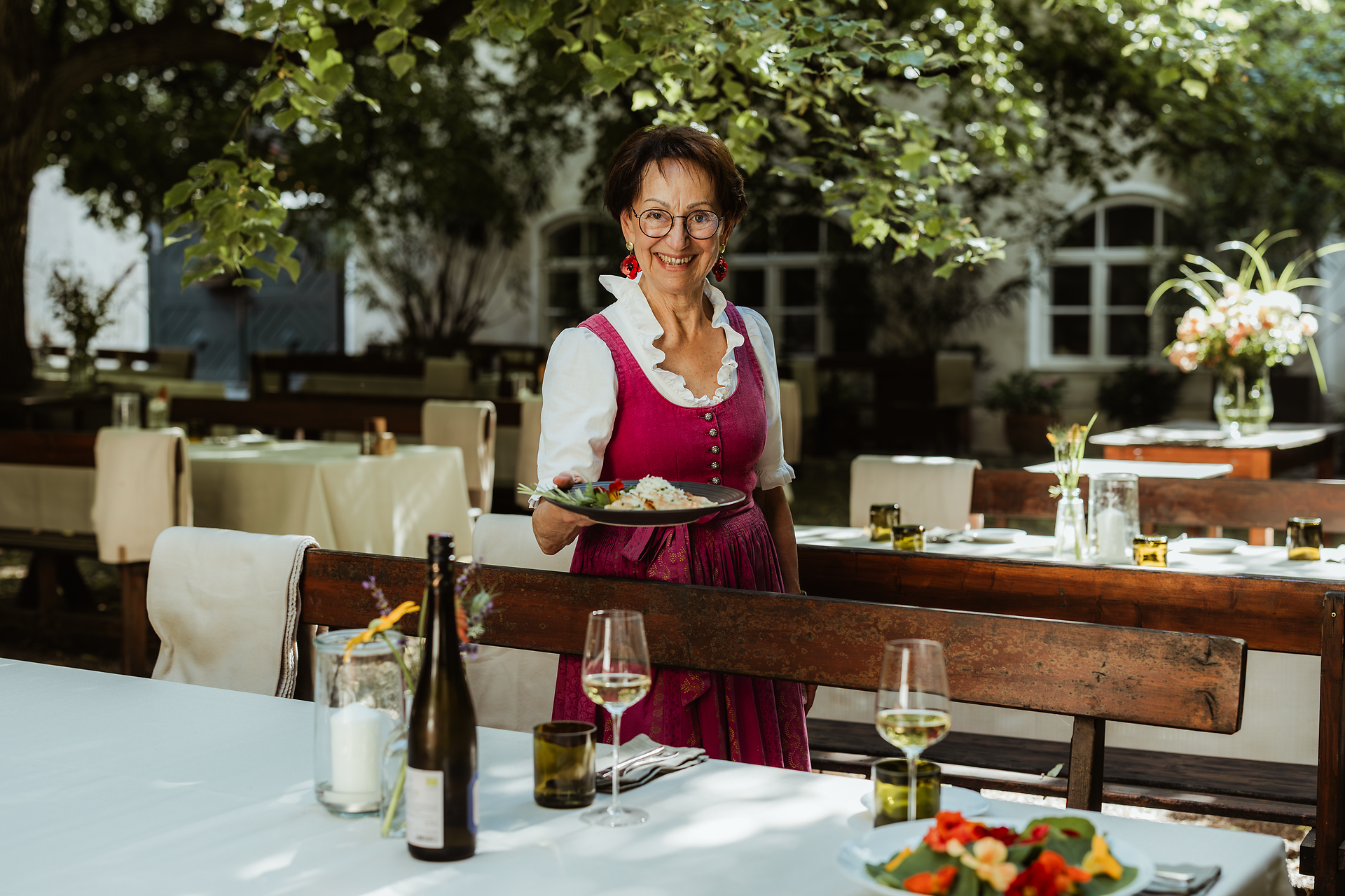 Eine Person im Dirndl steht in einem Gastgarten und hält einen Teller mit Speisen. Im Vordergrund sind ein gedeckter Tisch mit Weinflasche und Gläsern sowie Holzbänke zu sehen.