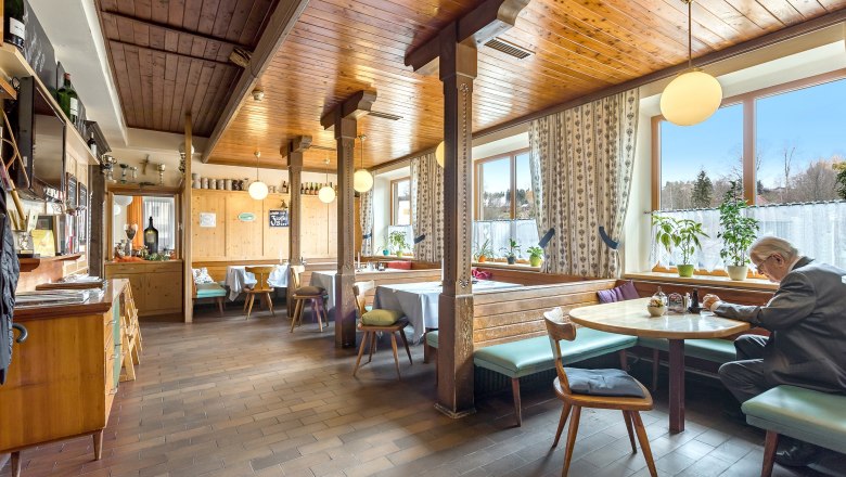 Gem&uuml;tliche Gaststube mit Holzm&ouml;beln und gro&szlig;en Fenstern, ein Mann sitzt an einem Tisch.