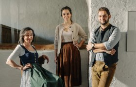 Drei Personen in traditioneller Kleidung in einem rustikalen Raum.