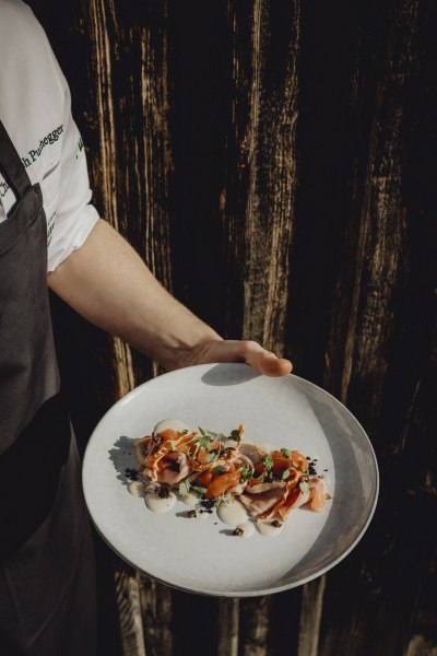 Ein Teller mit kunstvoll angerichtetem Vitello Forello, gehalten von einer Person in Kochkleidung vor einem Holzhintergrund.