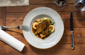 Ein Teller mit geschmorten Rindsbackerl, Ravioli und Gemüse auf einem Holztisch, daneben Besteck und ein Glas Rotwein.