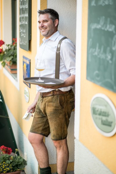 Ein Mann in traditioneller Kleidung lehnt an einer Wand und hält ein Tablett mit einem Glas Bier.